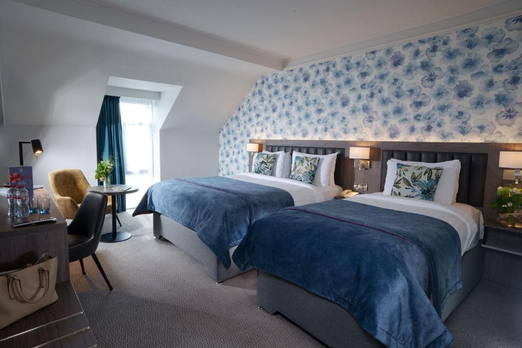 A hotel room with two beds with blue covers against a wall with blue floral wallpaper.