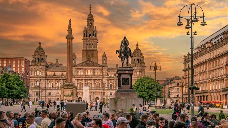 A bustling city square with people gathered in front of grand, historic buildings under a colorful sunset.