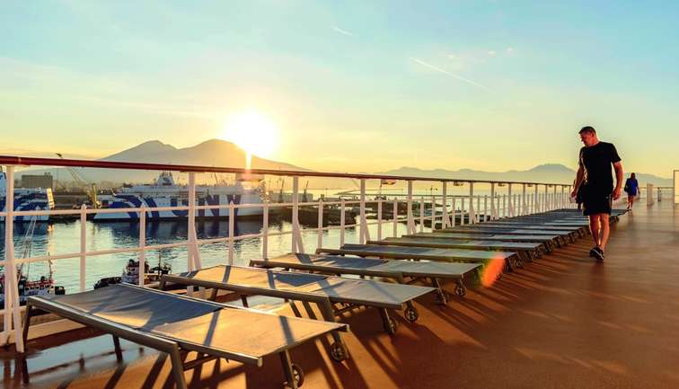 Man walking on a cruise ship deck at sunrise with mountains in the background.