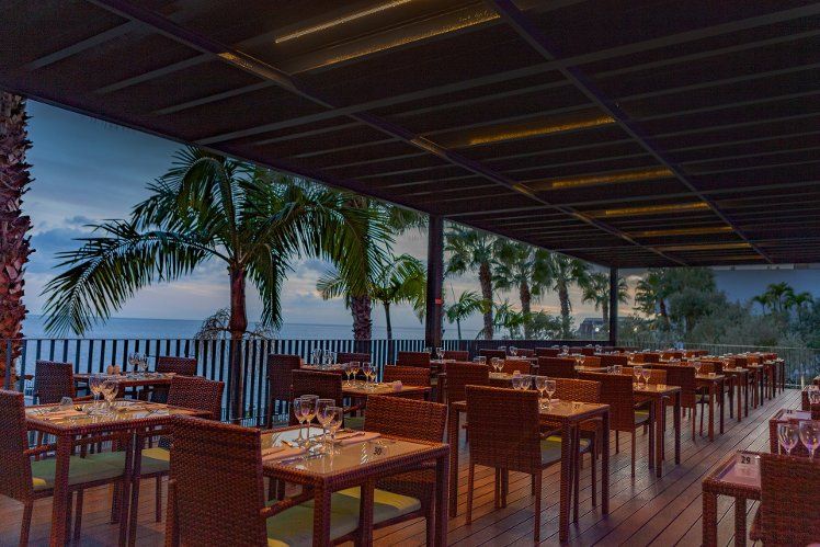 A covered outdoor restaurant patio with tables set for dinner, overlooking the ocean and palm trees at dusk.
