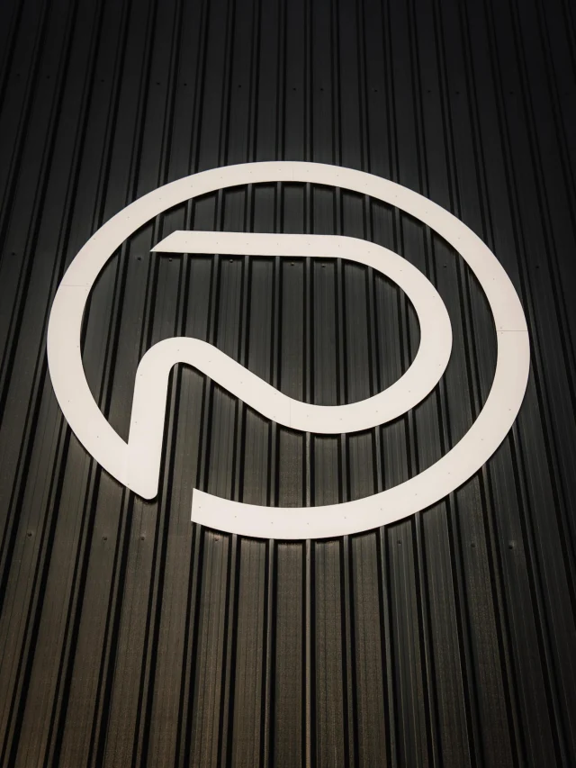 A white circular logo with a stylized letter P mounted on a black corrugated metal wall.