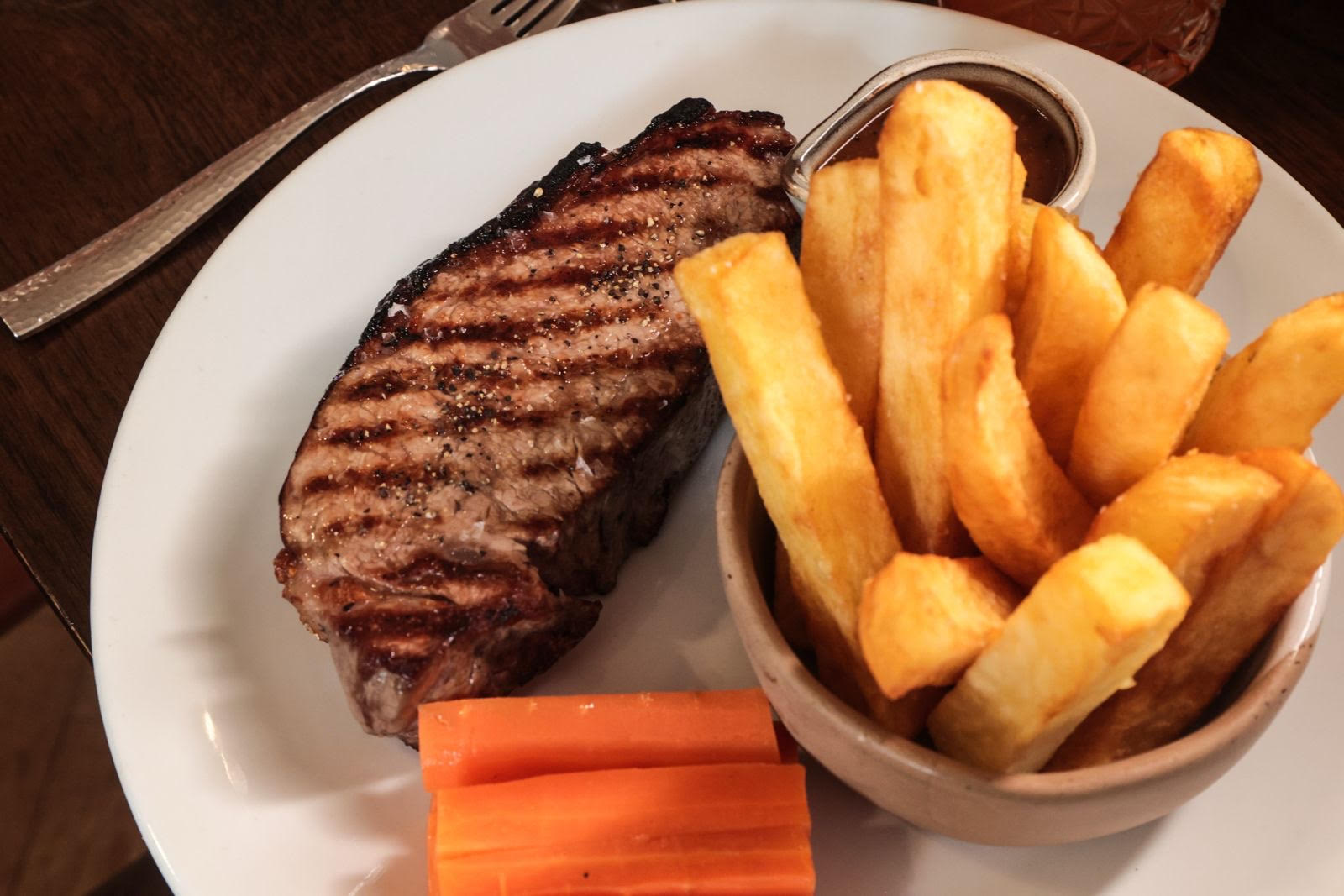 A grilled steak served with thick-cut fries, carrots, and sauce on a white plate.