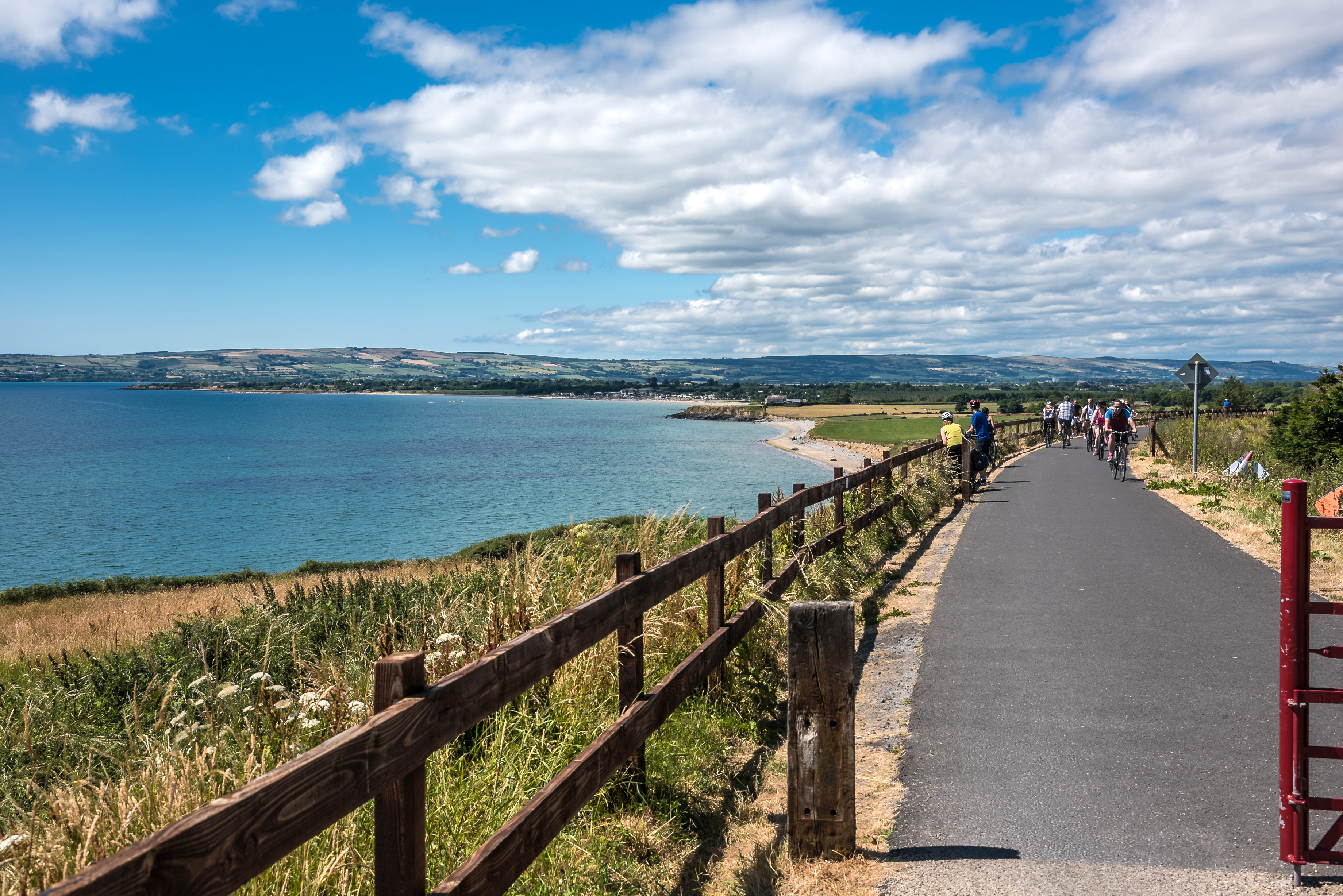 A group of cyclists rides on a paved path along a coastline on a partly cloudy day.