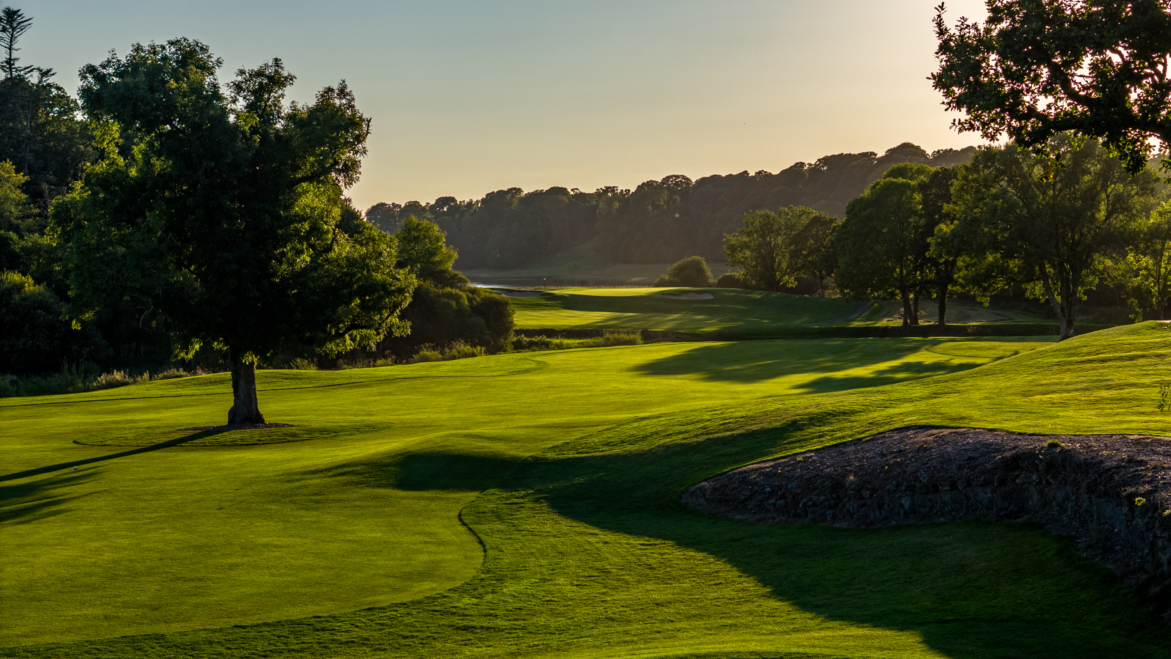 A scenic golf course with rolling green hills and trees bathed in warm, late afternoon sunlight.