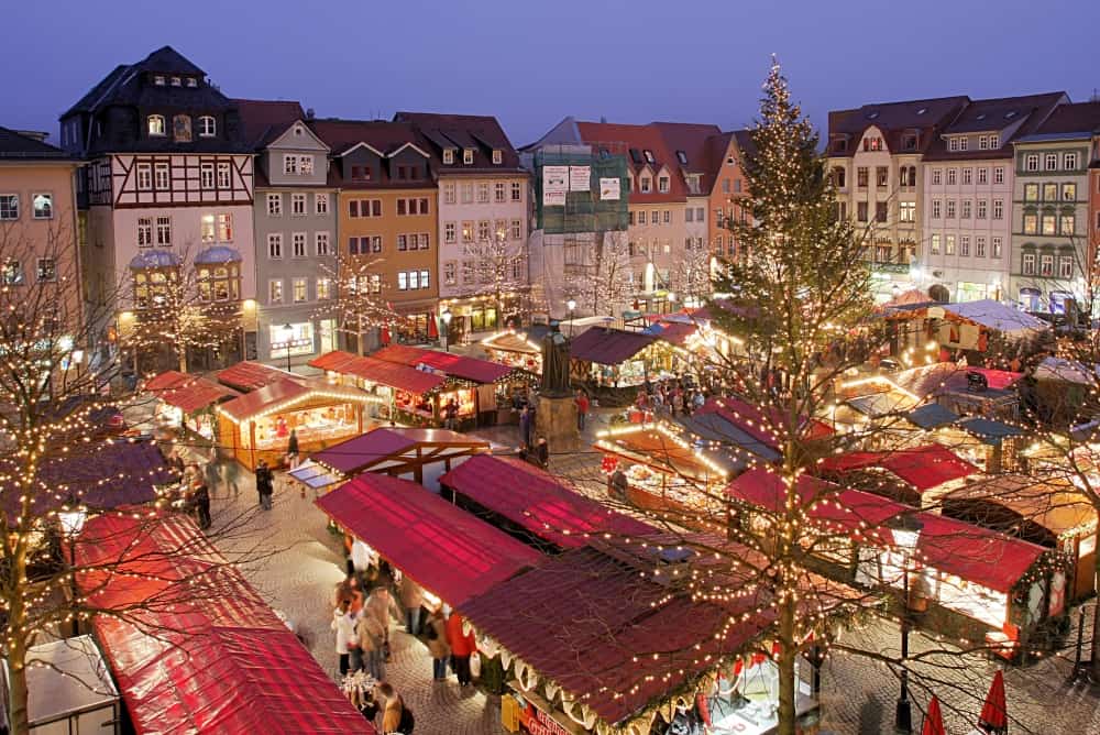 Festive market square with illuminated stalls and a large Christmas tree at dusk.
