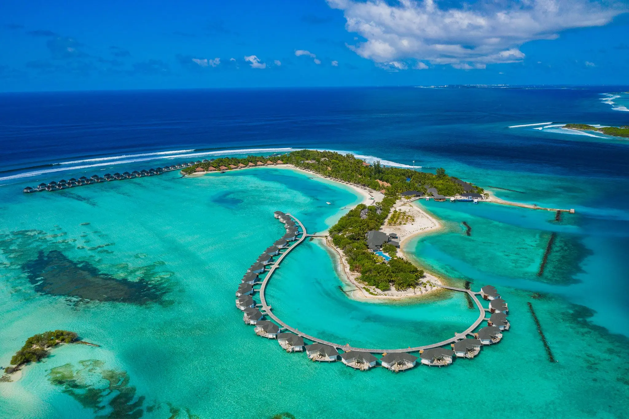 An aerial view of a tropical island resort with overwater bungalows arranged in a curve over turquoise water.