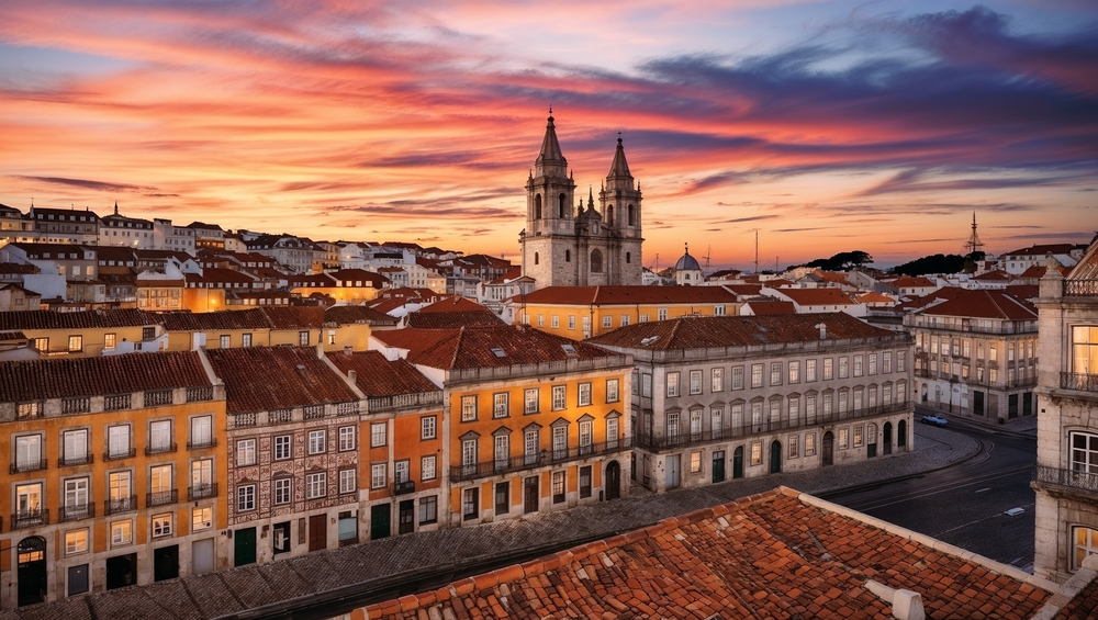 A cityscape with red-tiled roofs and a prominent church with two towers under a colorful sunset.