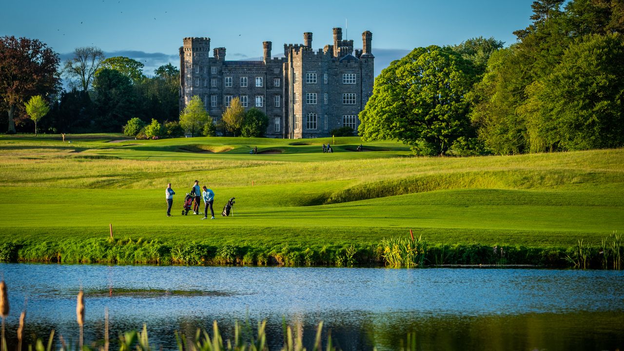 Golfers on a lush green course with a large stone castle and a pond.