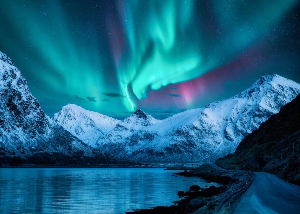 The aurora borealis shines over snow-covered mountains, a body of water, and a road at night.