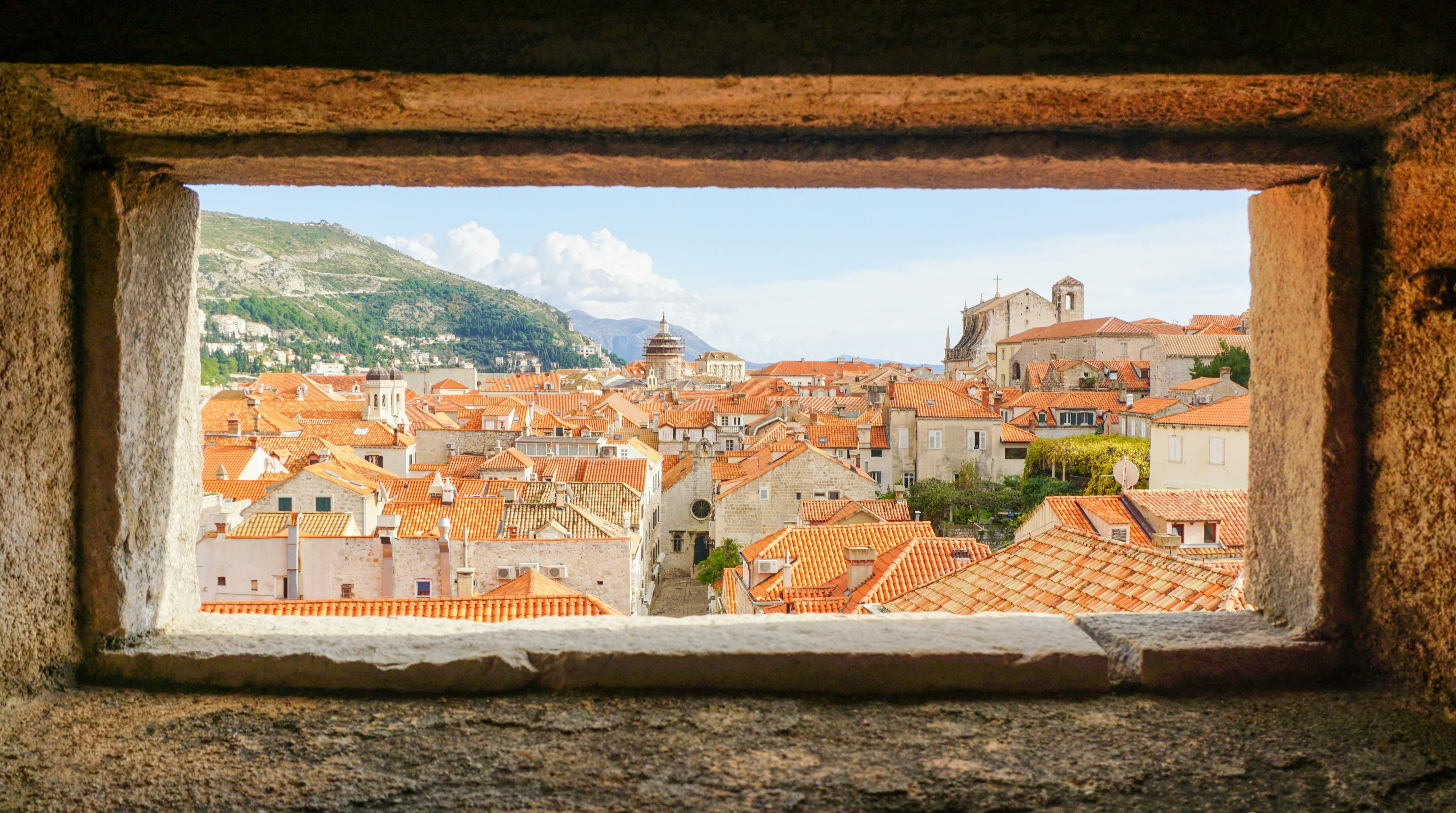 View of a city with red-tiled roofs through a stone window frame.