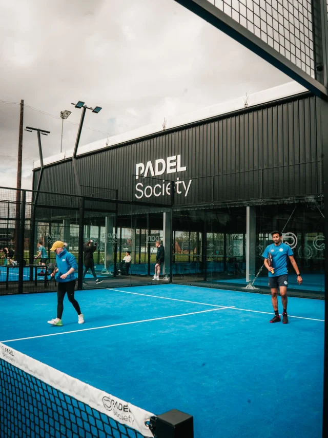 People playing on a bright blue padel court in front of the Padel Society building.
