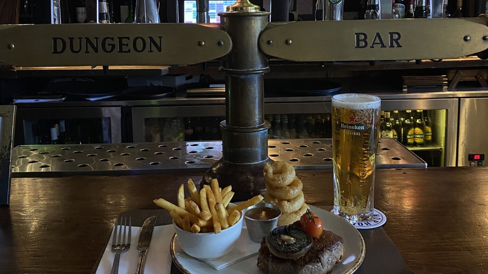 A meal of steak, fries, and onion rings with a glass of beer on a bar counter.