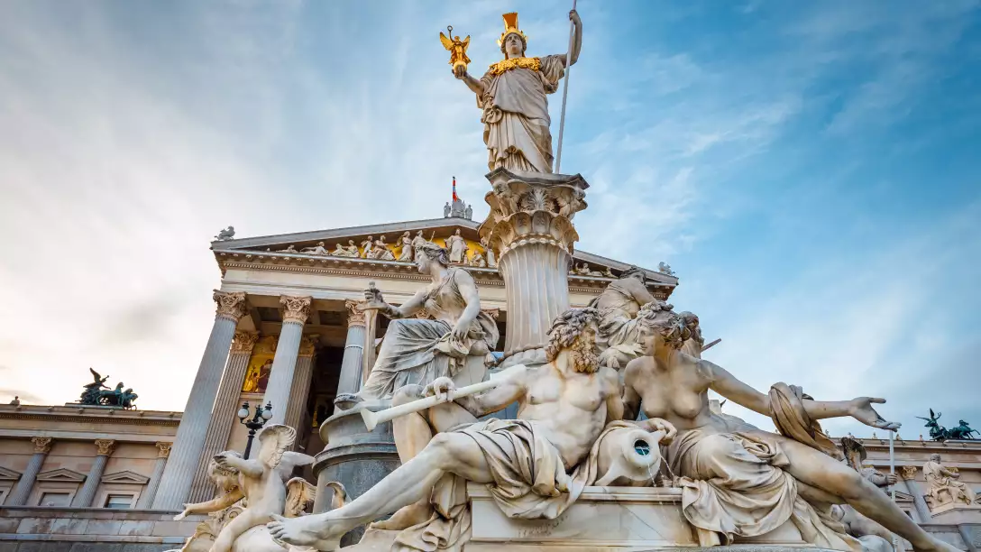 A marble fountain with classical statues in front of a grand building with columns under a partly cloudy sky.