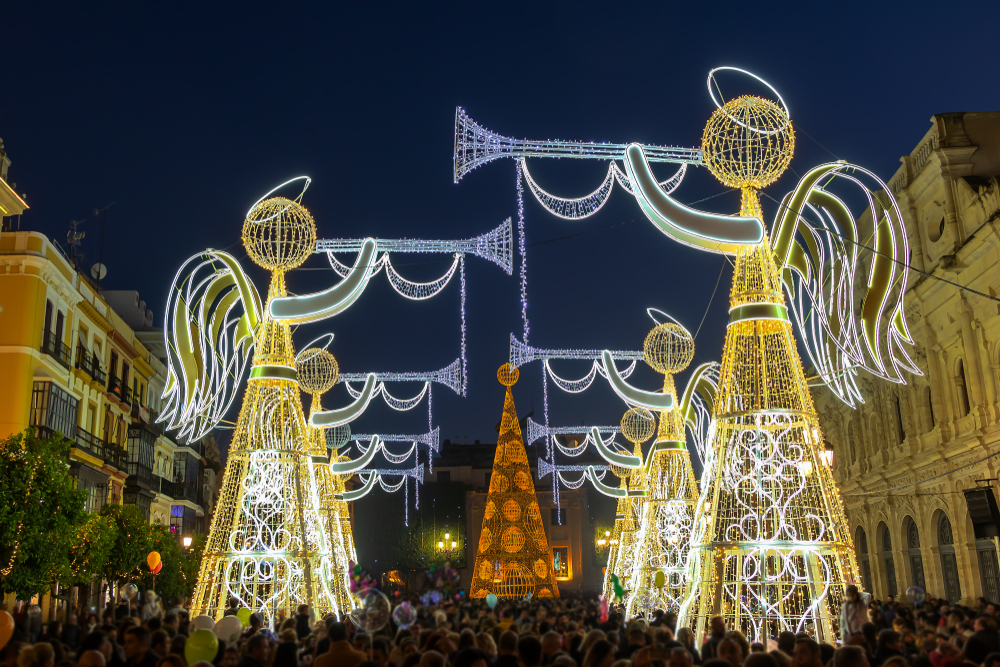 A crowd watches enormous, lit-up angel decorations holding trumpets over a street at night.