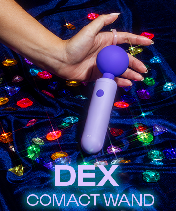 A hand holding a purple wand massager over a blue velvet cloth with colorful sparkling gems.