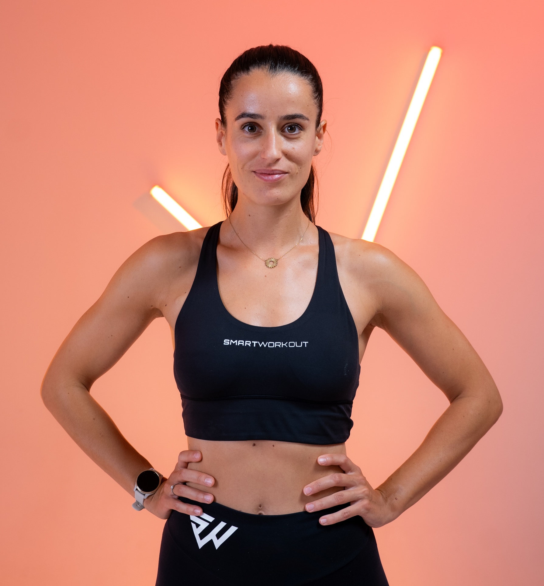 A woman in a black workout outfit stands with her hands on her hips against a pink background.