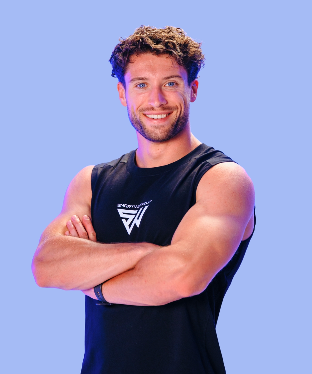 A smiling, fit man with curly hair and blue eyes, wearing a black tank top with his arms crossed.