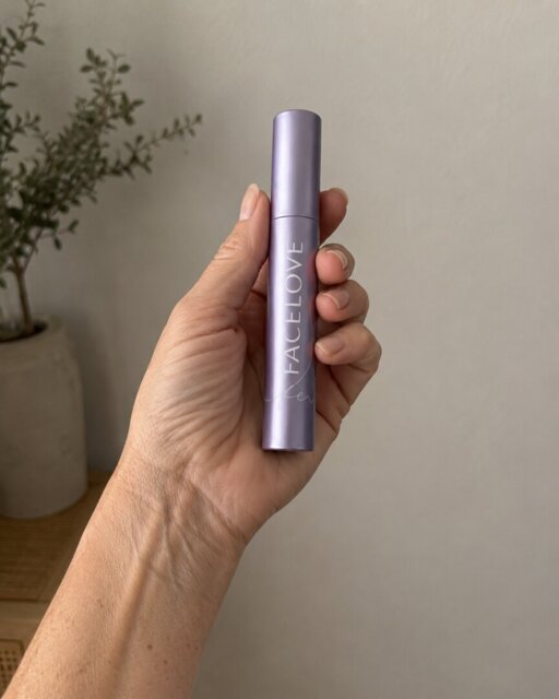 A hand holds a light purple tube of Facelove mascara against a plain wall with a plant.