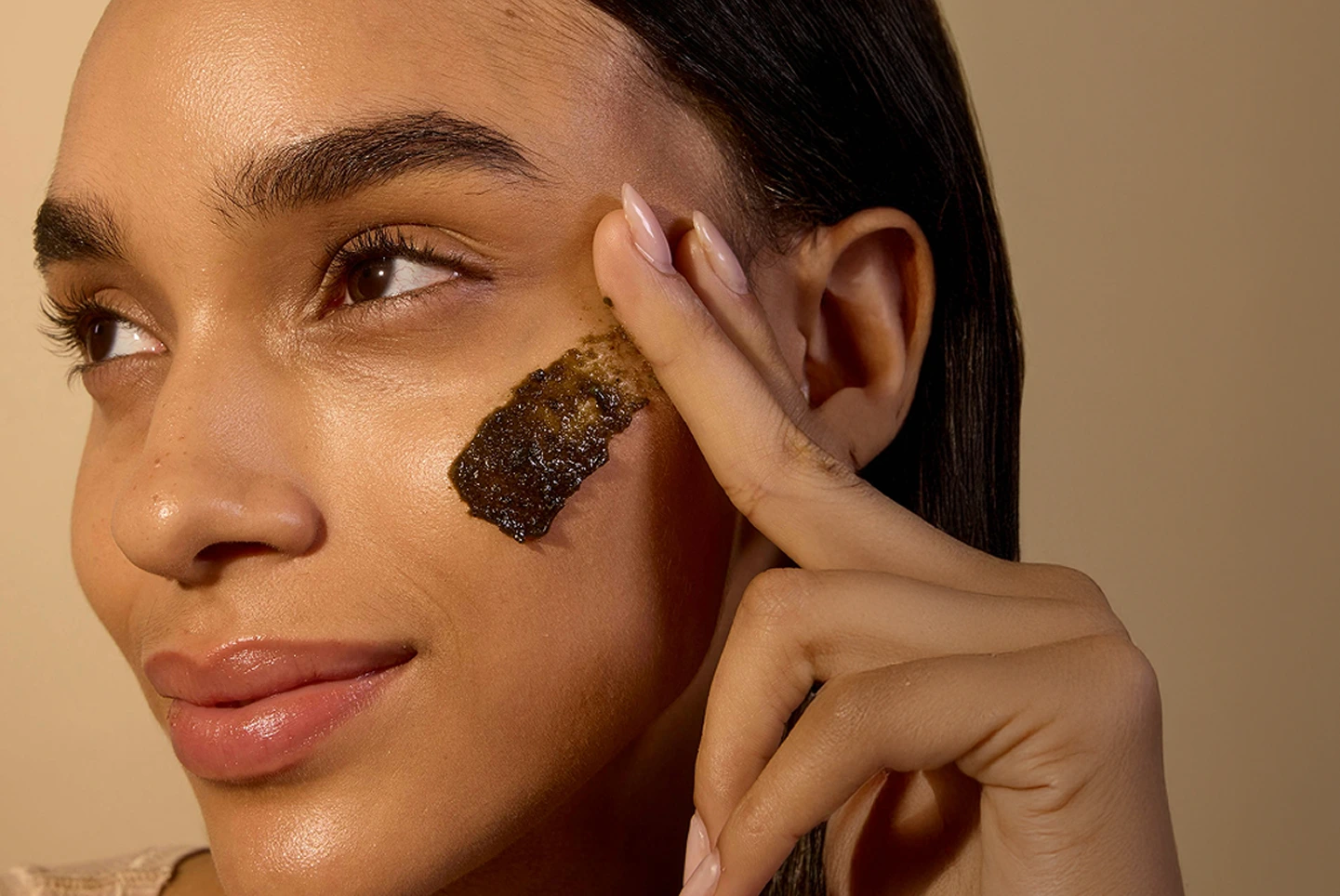 Close-up of a person applying a dark facial scrub on their cheek.