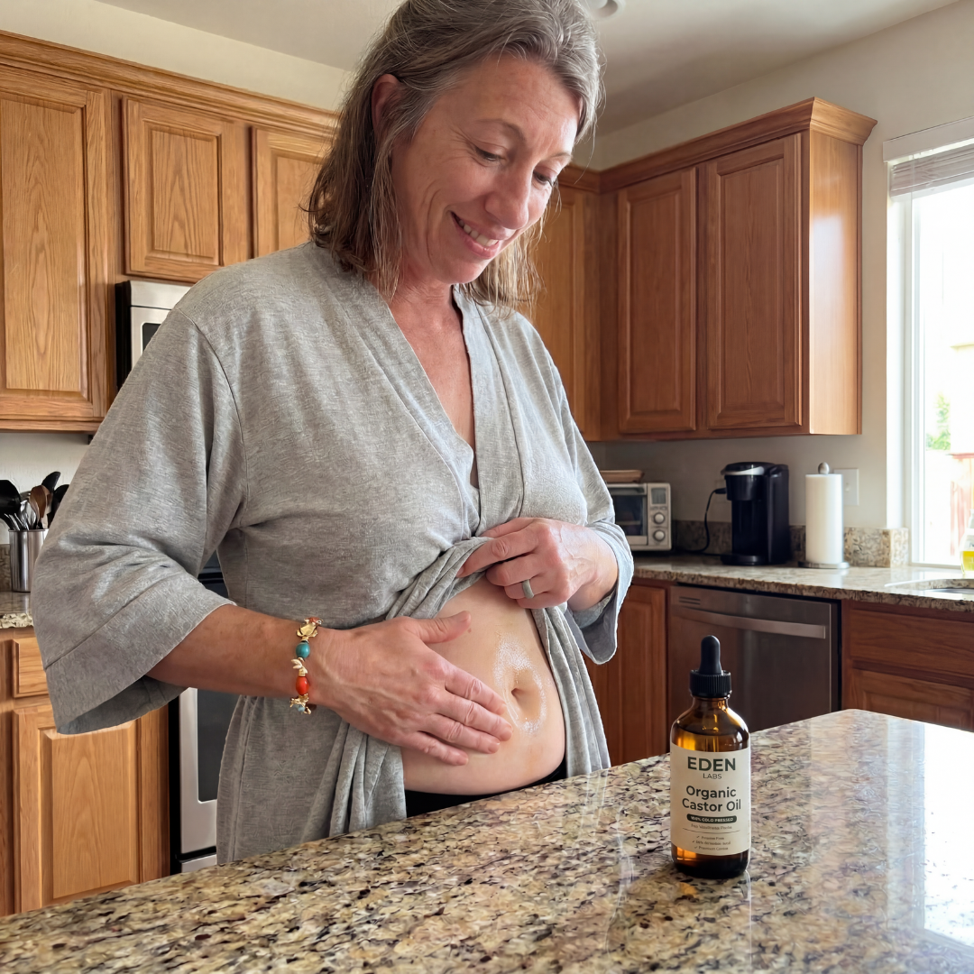 A woman in a grey robe smiles while applying oil to her stomach in a kitchen.