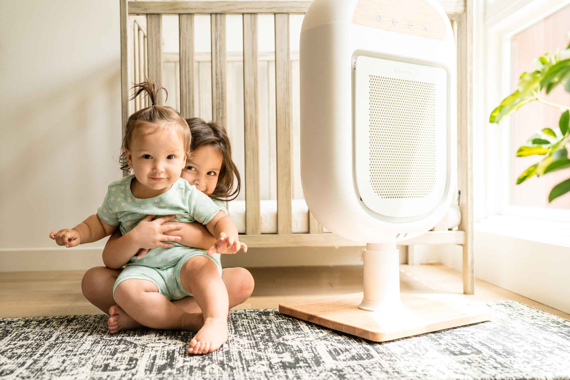 Two children sitting on the floor near a crib and air purifier.
