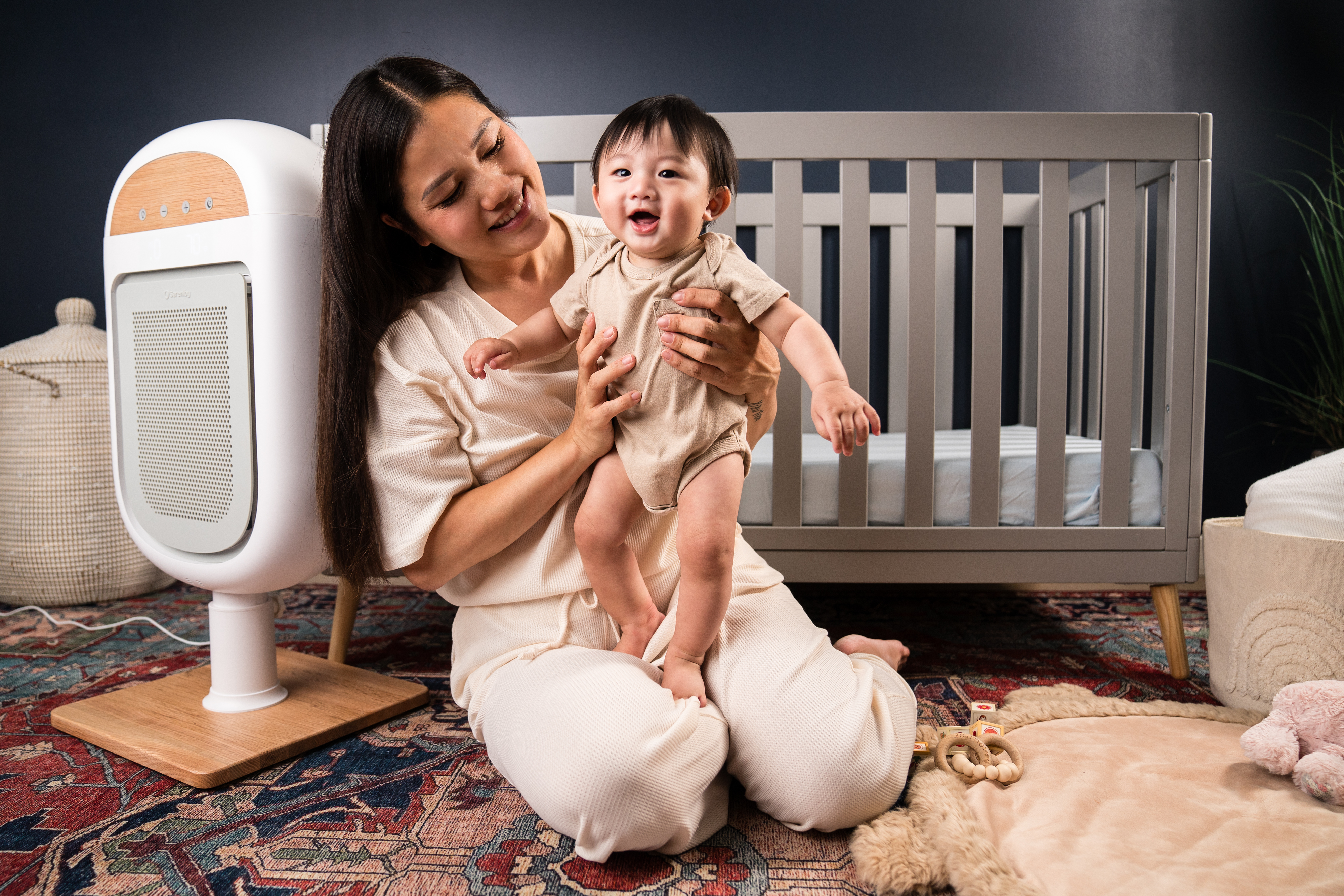 Woman holding a smiling baby in front of a crib and air purifier.