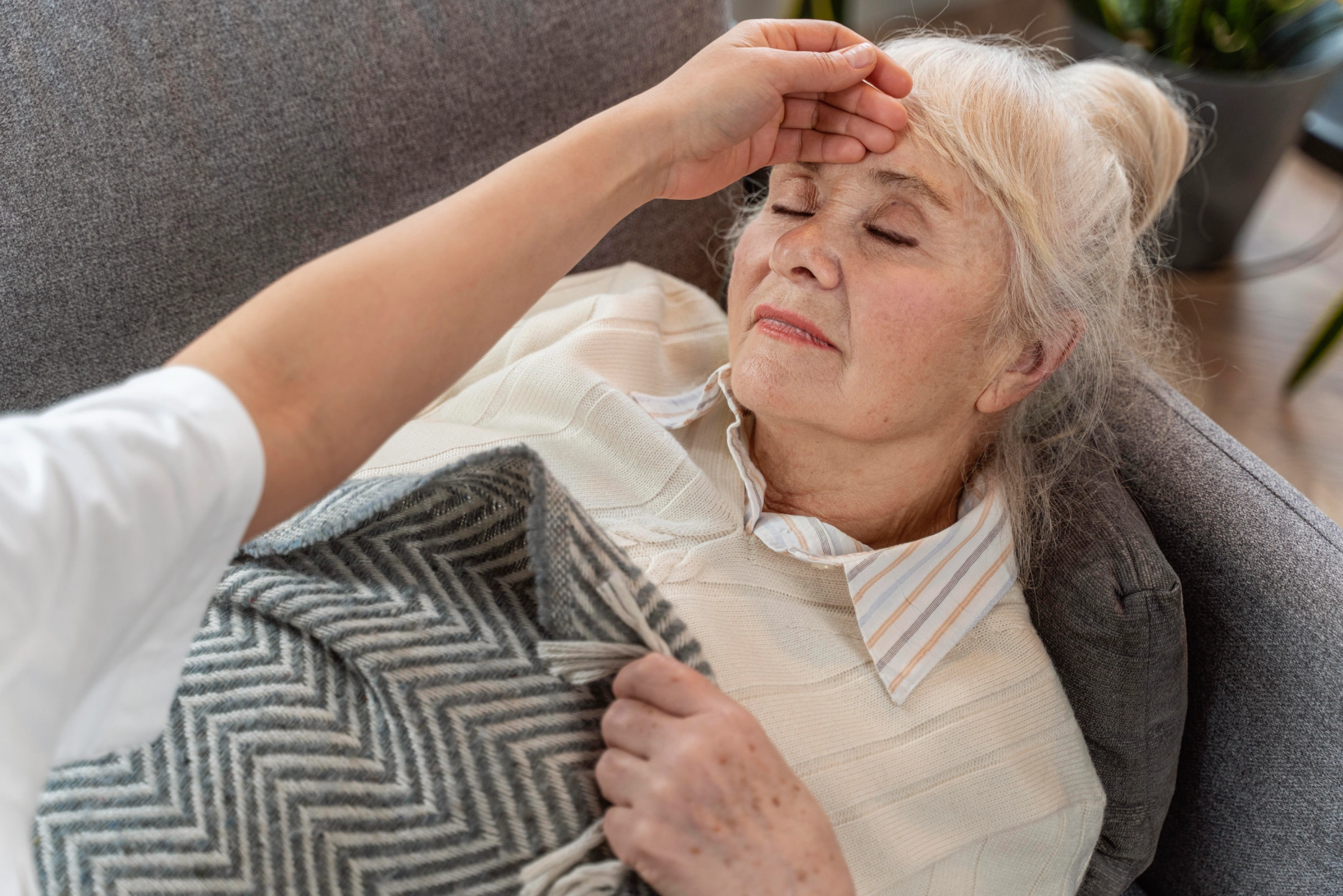 Elderly woman resting on a sofa with a hand on her forehead and holding a blanket.