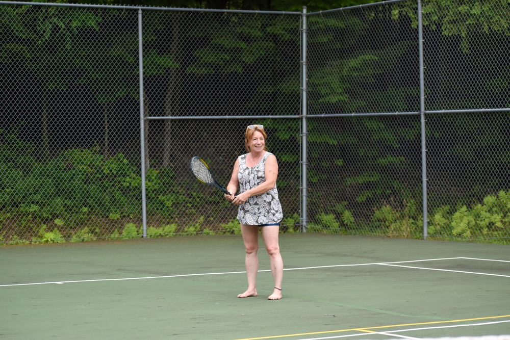 Woman standing on tennis court holding a racket, surrounded by greenery.