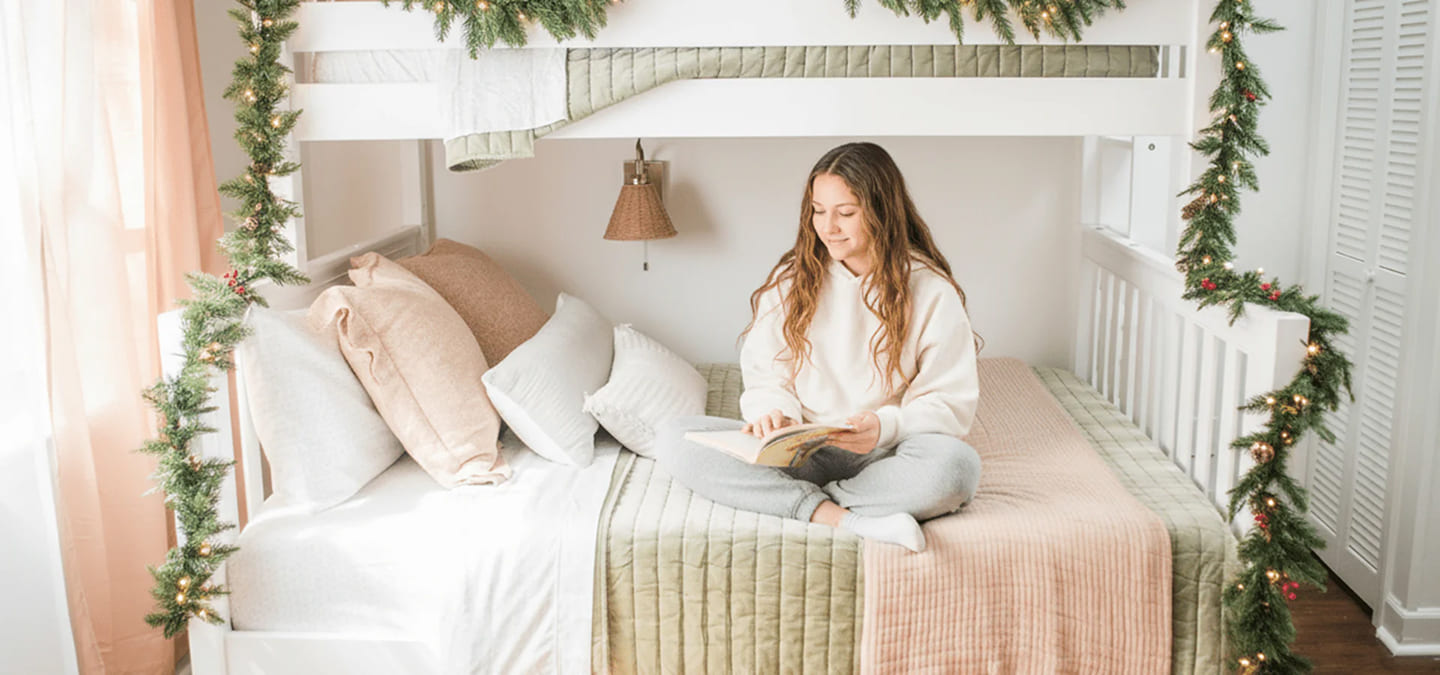 Teenage girl reading a book on her white bunk bed.