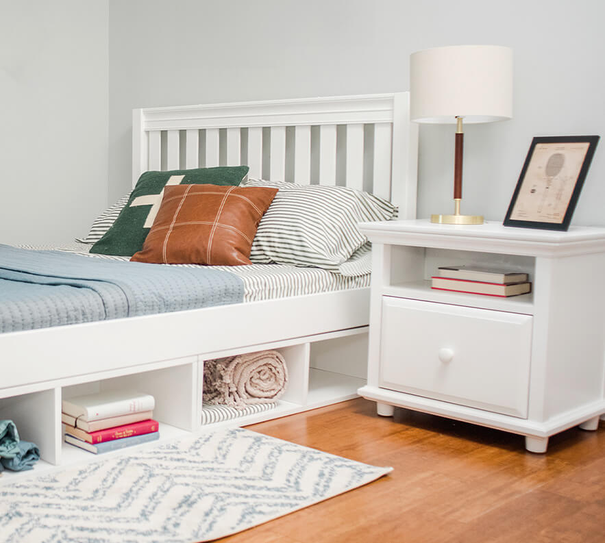 White wooden bed frame next to matching nightstand with lamp, books, and decorative pillows in modern bedroom.