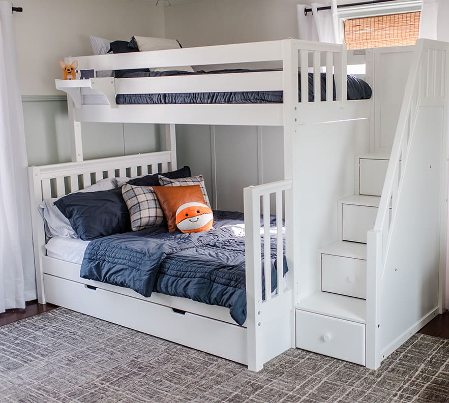 White wooden bunk bed with a cozy bottom bunk, blue bedding, a playful orange pillow, and built-in staircase drawers, perfect for kids' bedroom storage solutions.