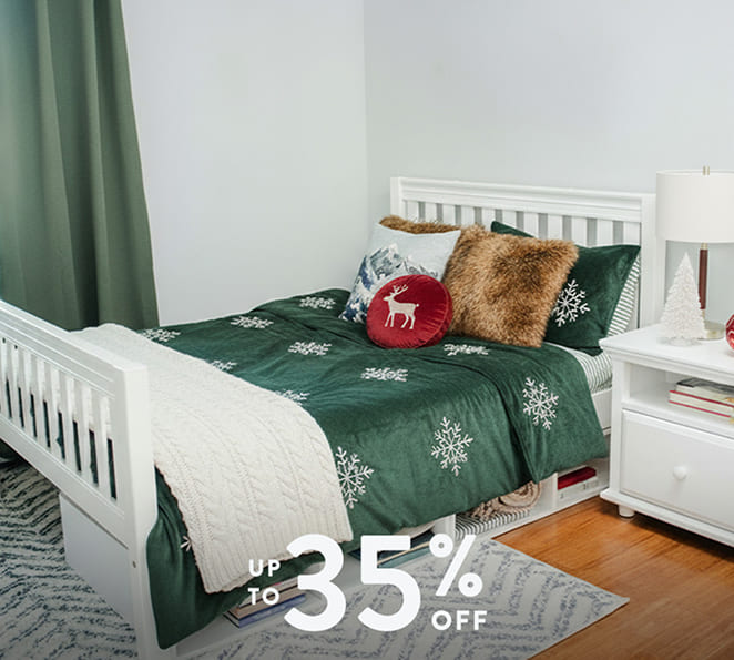 Green snowflake Christmas bedding set and red reindeer accent pillow on a white wooden bed with faux-fur and knit throws, white nightstand with lamp — holiday bedroom decor, up to 35% off.