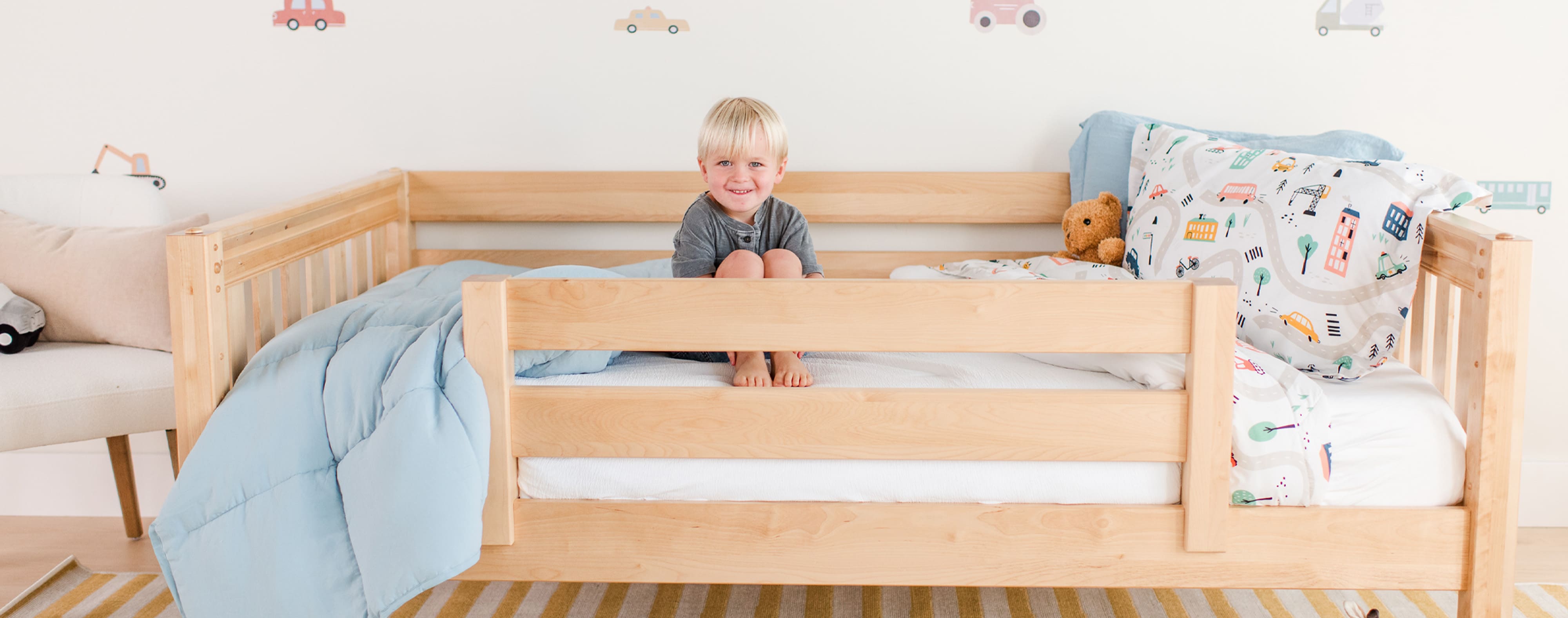 Toddler sitting in a wooden bed with colorful bedding and teddy bear in a cozy children's room.