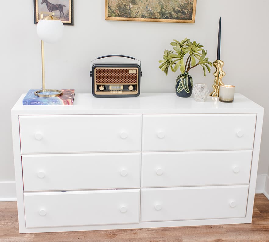 White dresser with vintage radio, lamp, plant, and candle on top in a modern interior setting.