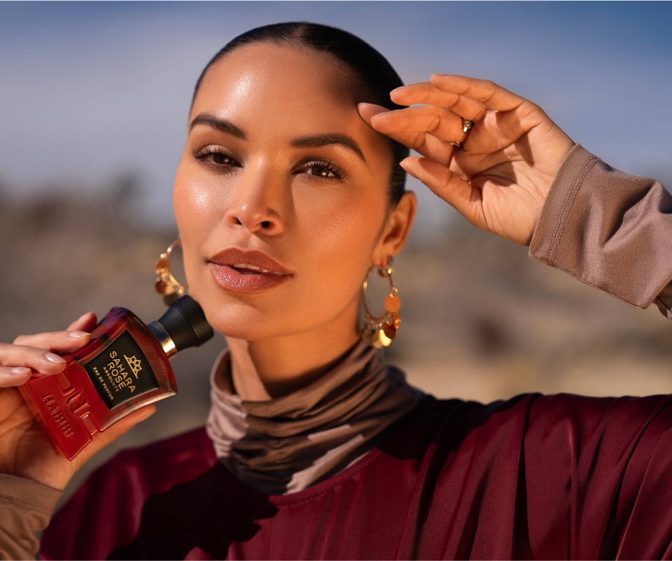 Person holding a perfume bottle outdoors, wearing earrings and a maroon outfit.