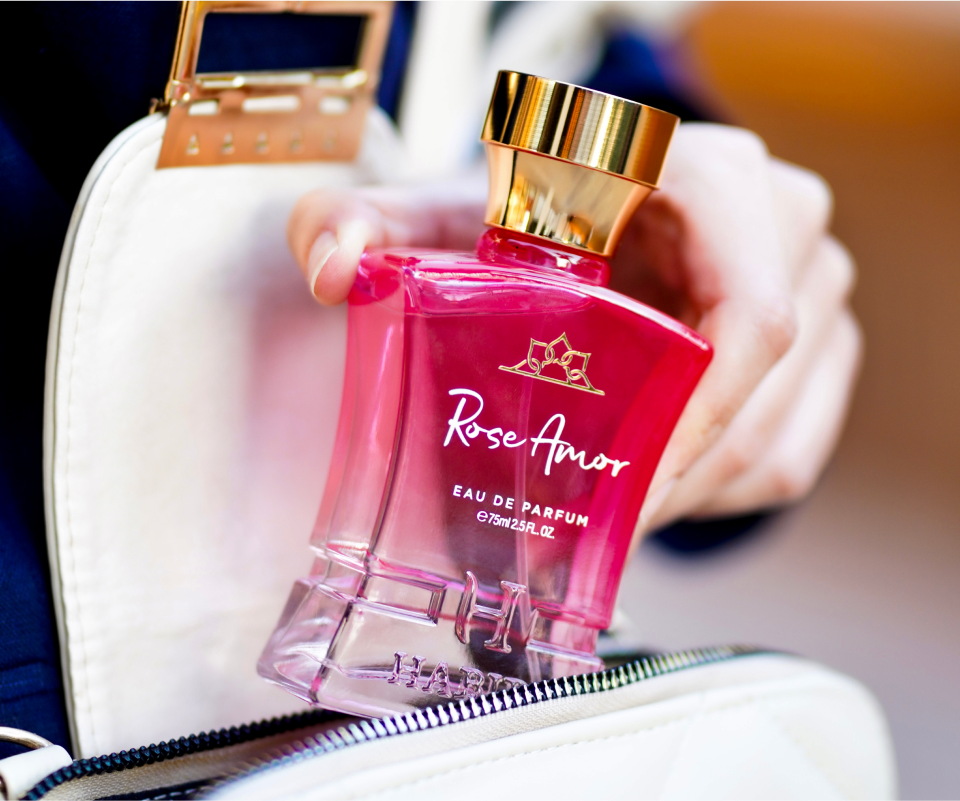 Hand holding a pink perfume bottle labeled 'Rose Amor' near a white bag.