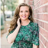 Woman in a green dress smiling, leaning against a brick wall.