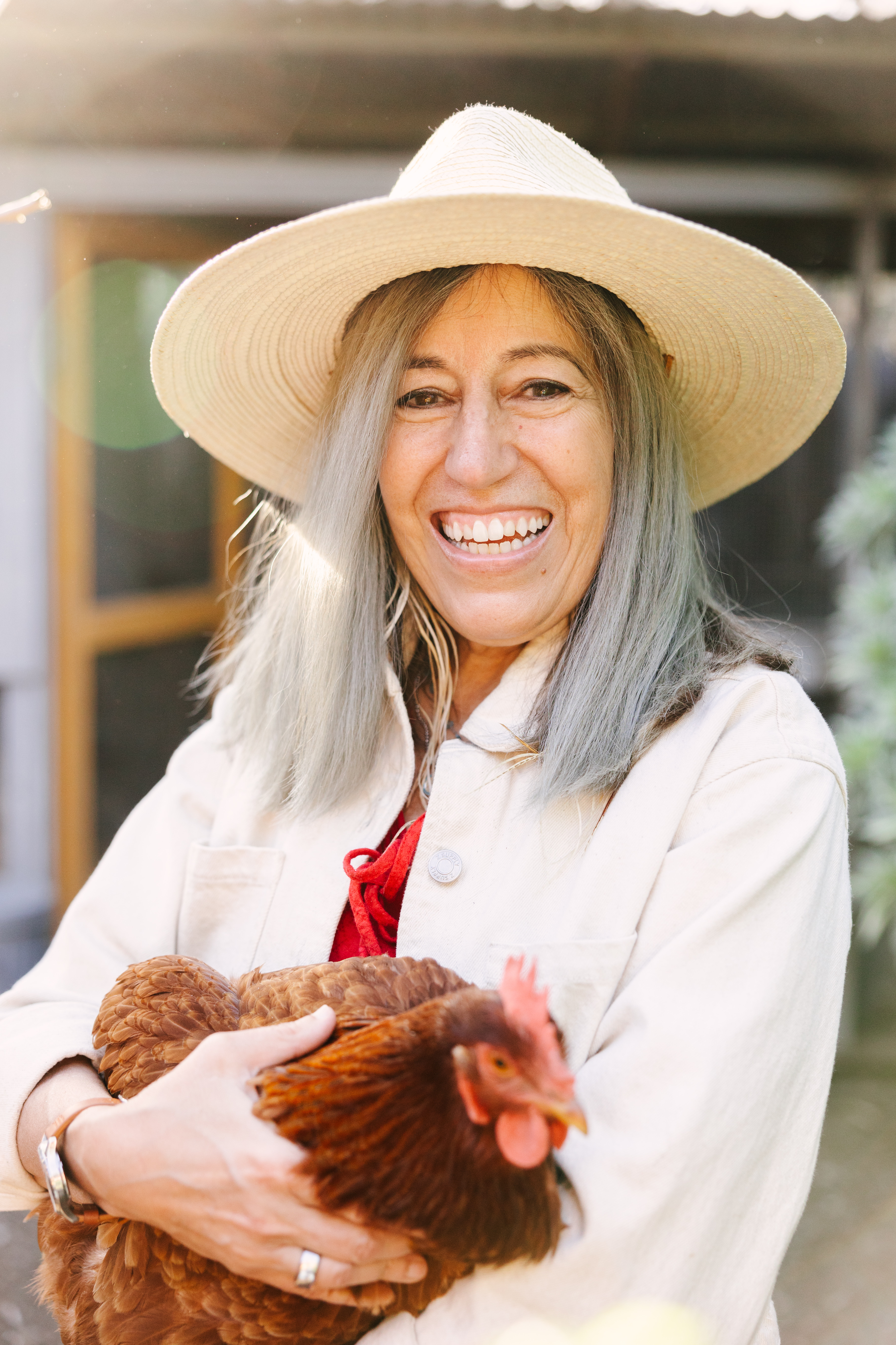 A smiling woman with gray hair wearing a straw hat holds a brown chicken in her arms.