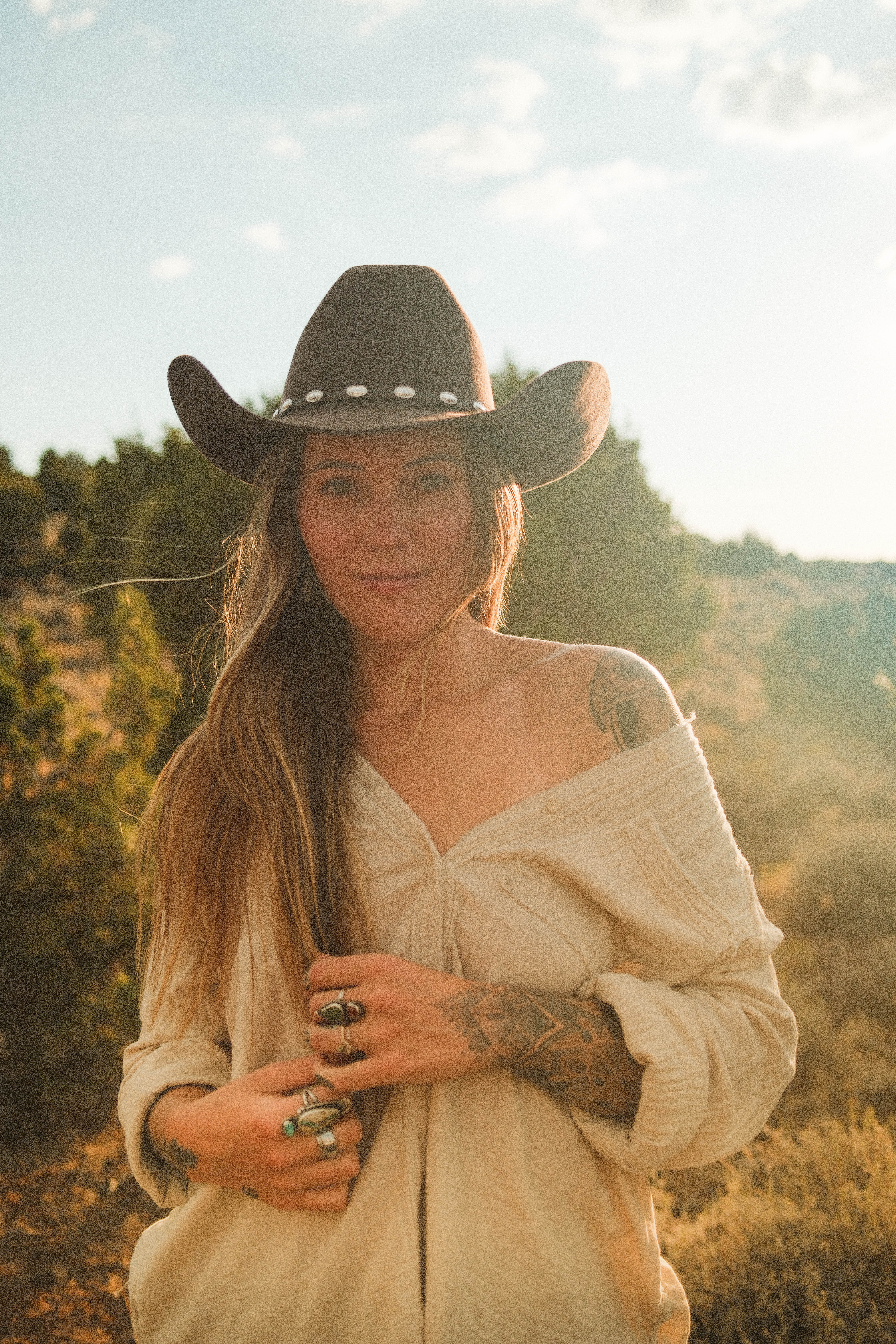 A woman with tattoos and long hair wearing a studded cowboy hat outdoors during golden hour.