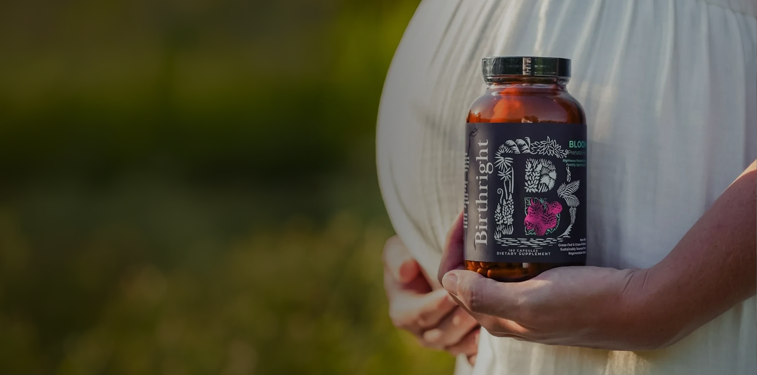 A pregnant person in a white dress holds a bottle of Birthright prenatal supplements against their belly.
