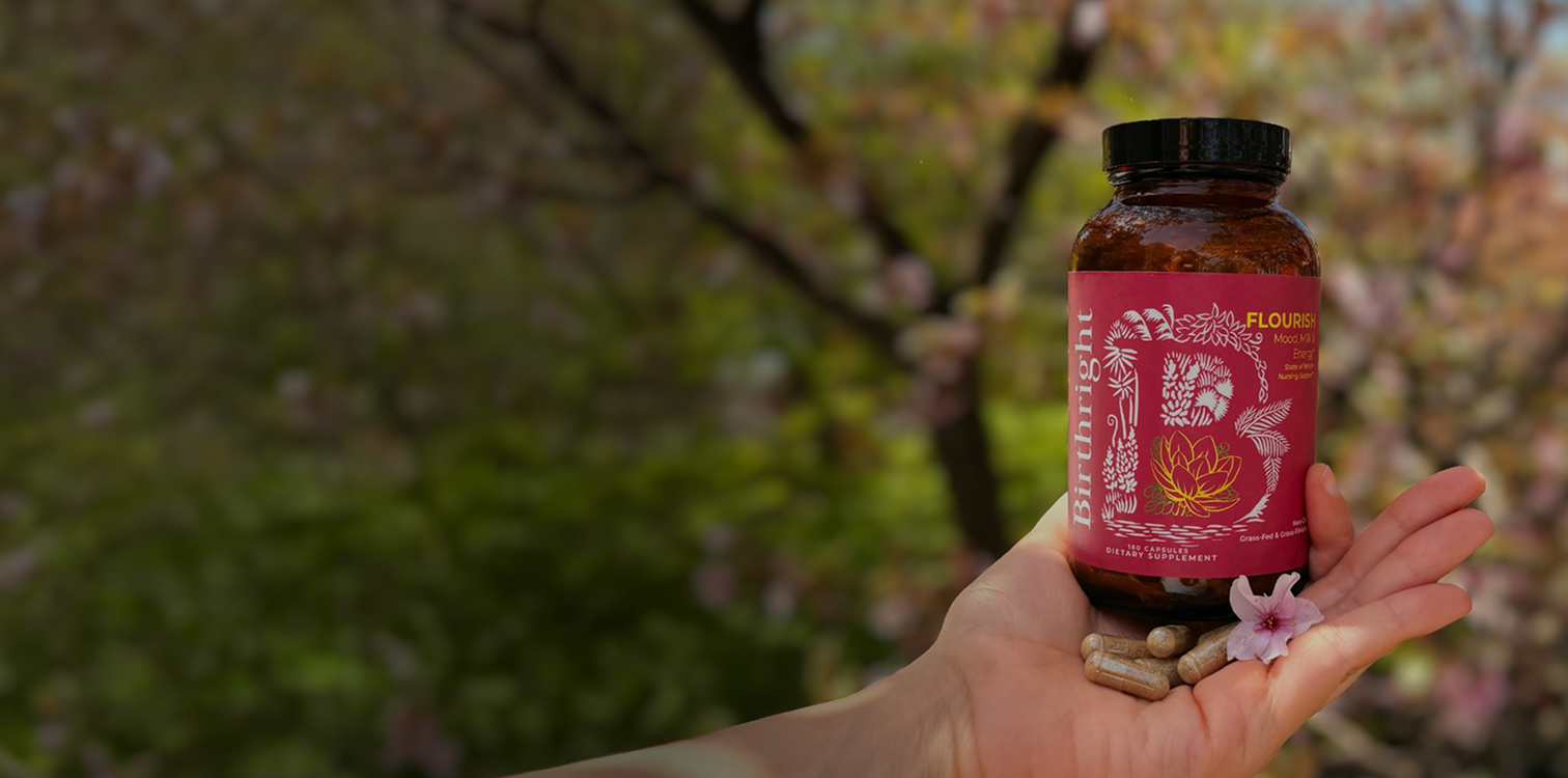 A hand holds a bottle of Birthright Flourish dietary supplements, some capsules, and a pink flower outdoors.