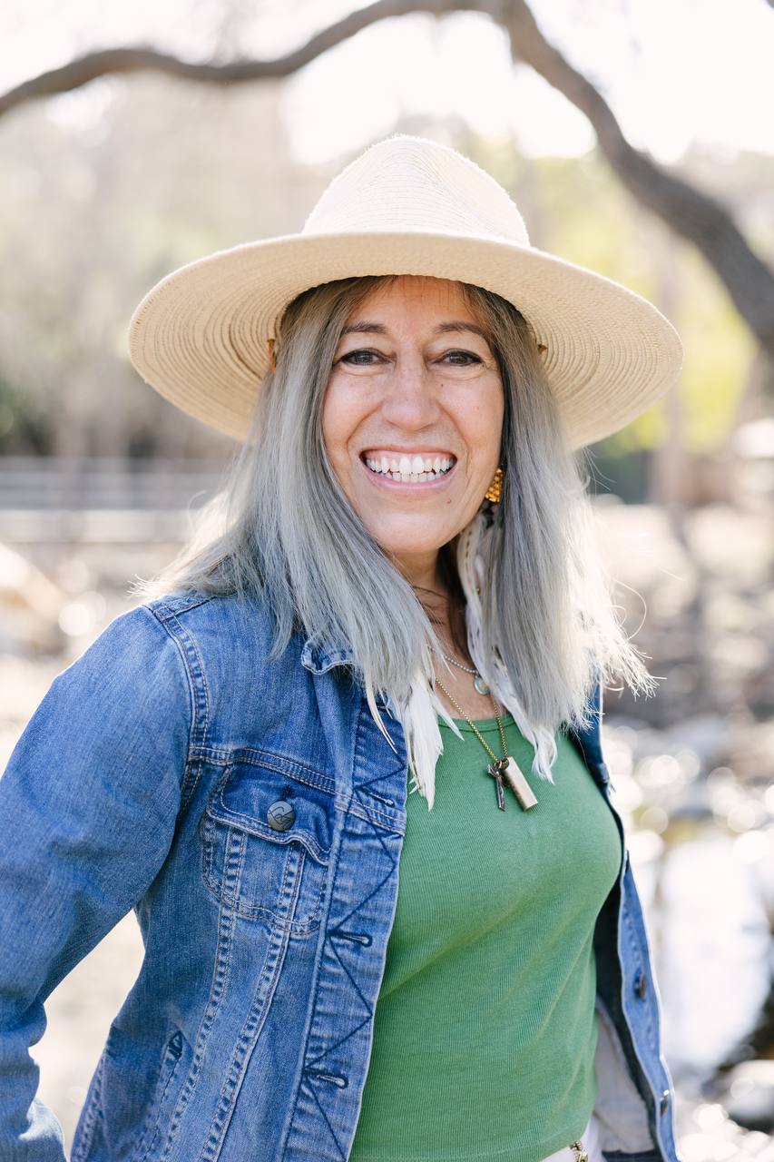 A smiling woman with grey hair, wearing a straw hat and denim jacket, stands outdoors.