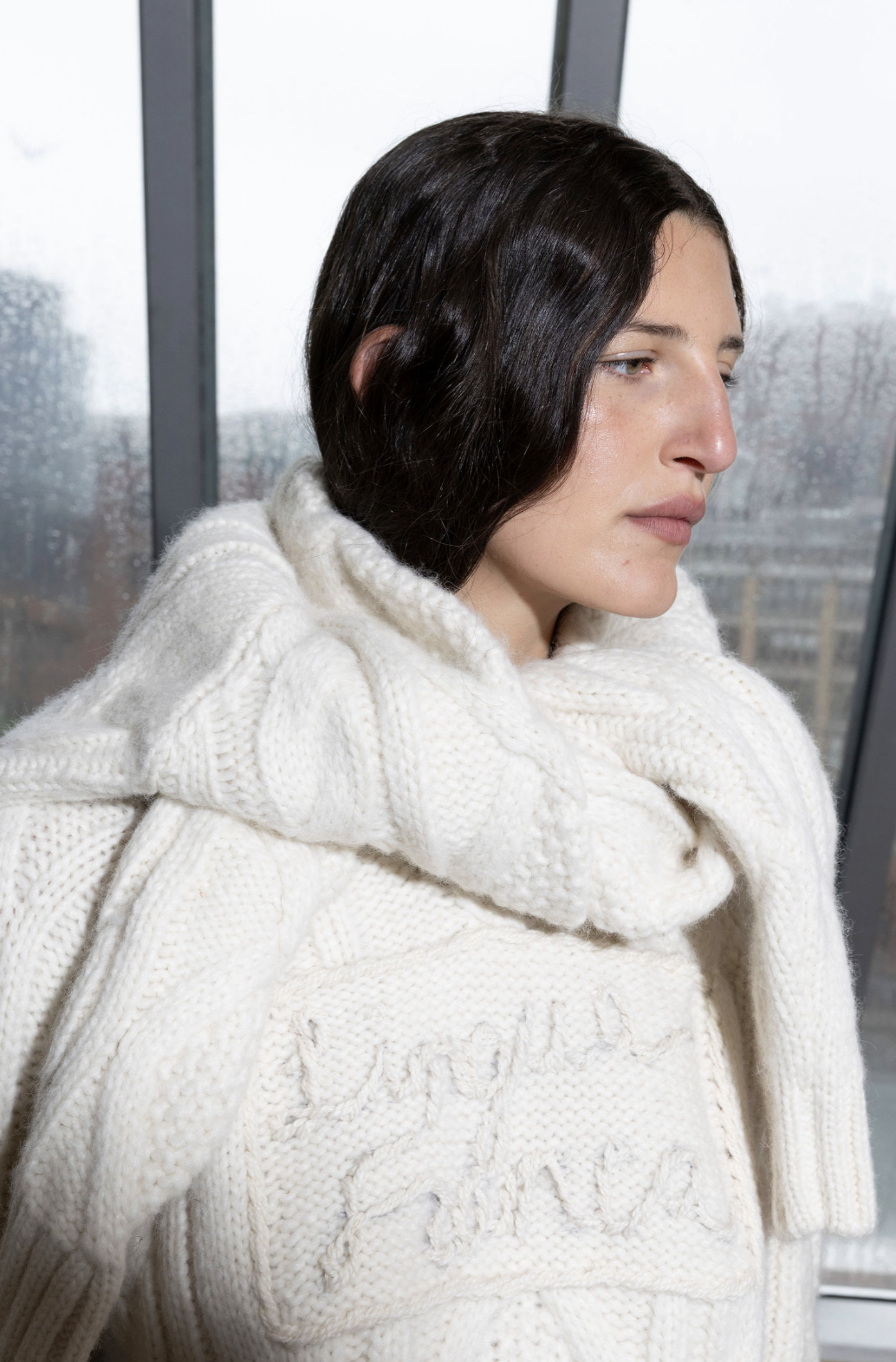 Person wearing a white knitted sweater and scarf near a window on a rainy day.