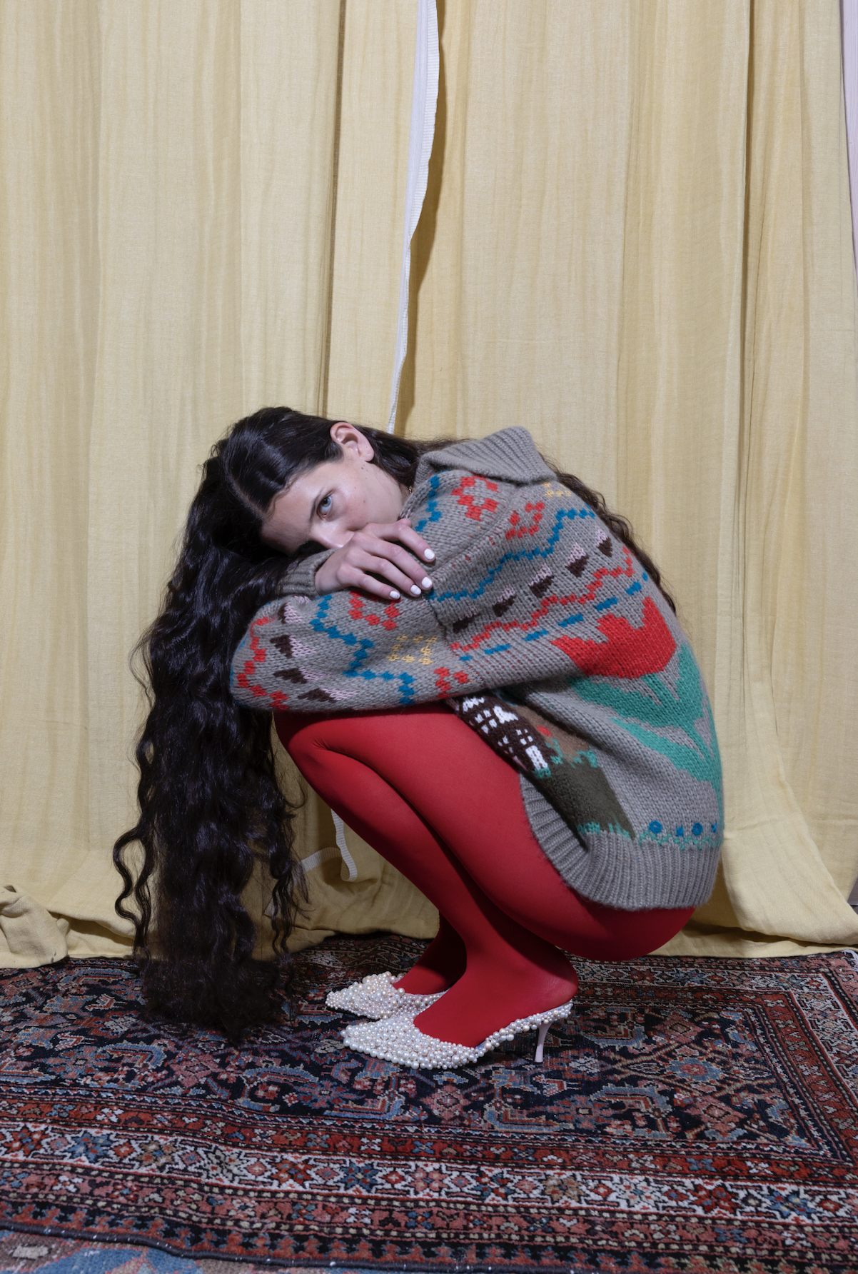 Person crouched on a rug, wearing a colorful sweater and red tights with pearl shoes.