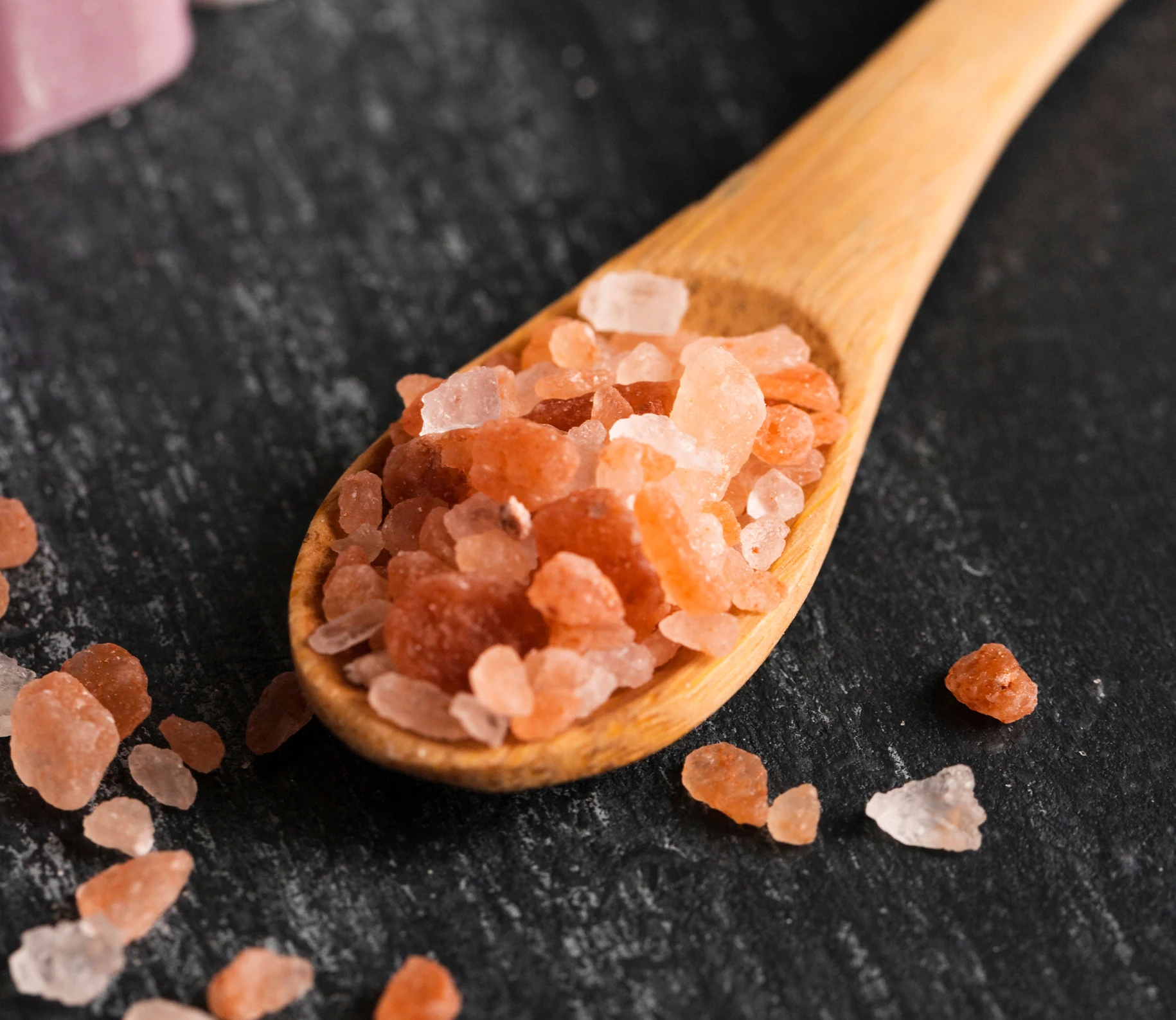 A wooden spoon with pink rock salt on a dark surface.