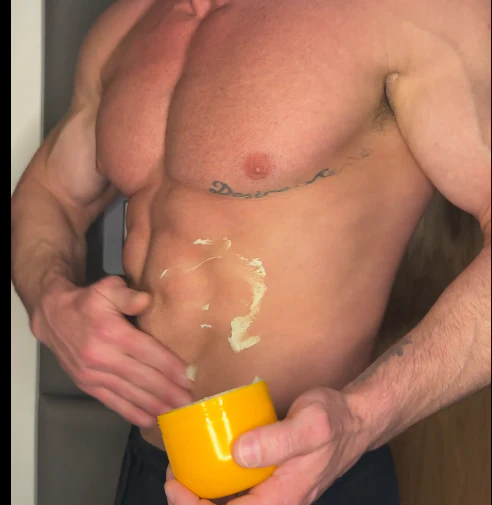 A muscular man with a chest tattoo applies cream from a yellow jar to his abdomen.