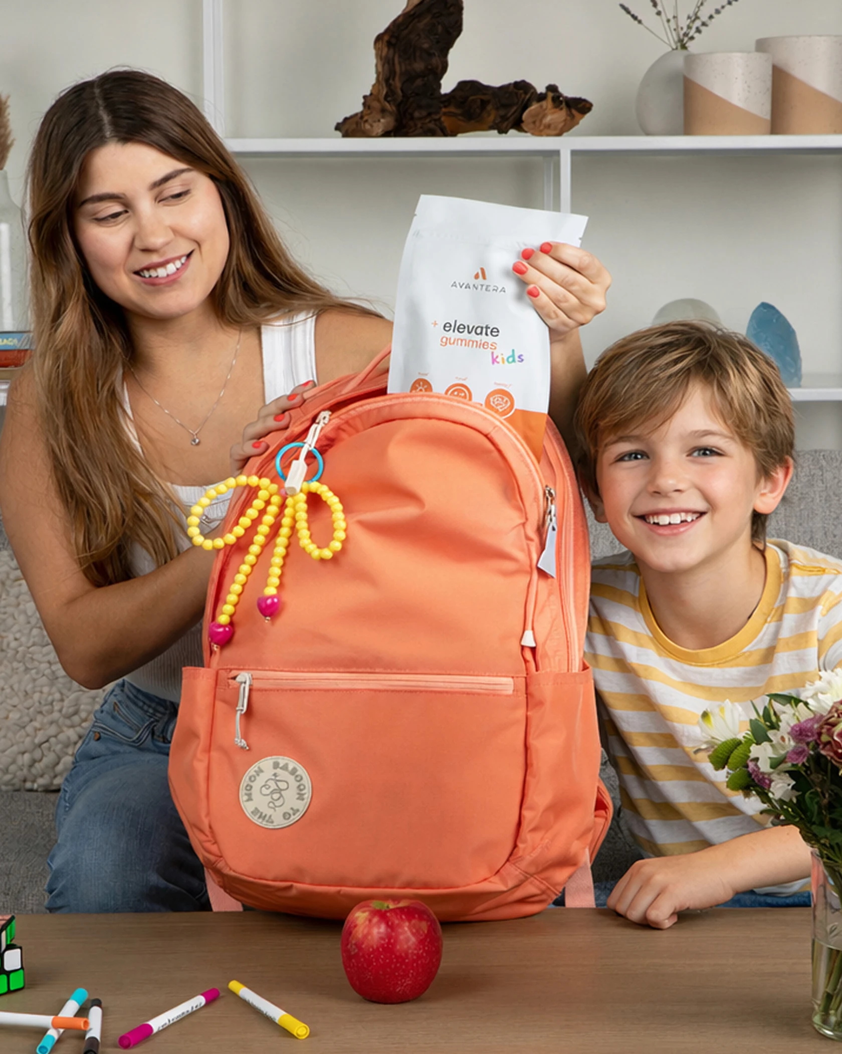 A woman places a bag of children's gummies into an orange backpack next to a smiling boy.