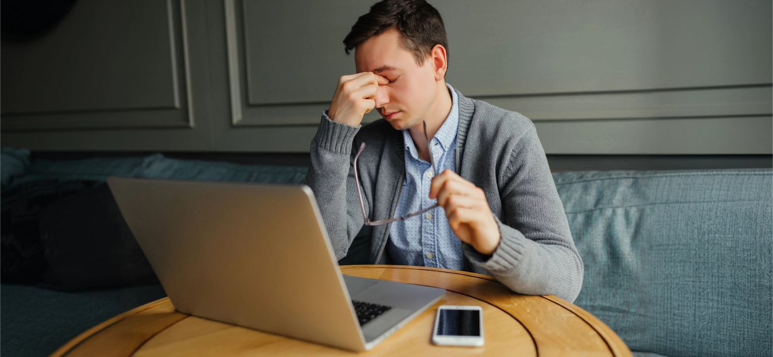 A stressed-looking man pinches the bridge of his nose while sitting at a table with a laptop.