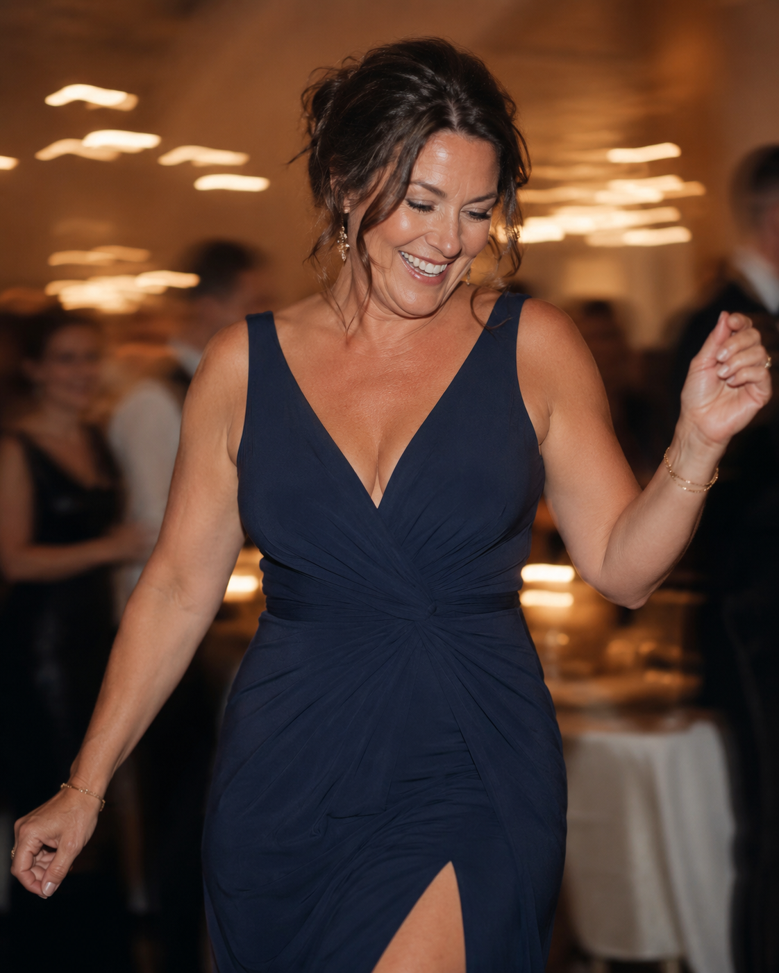 A smiling woman in a dark blue, v-neck gown with a side-slit is dancing indoors.