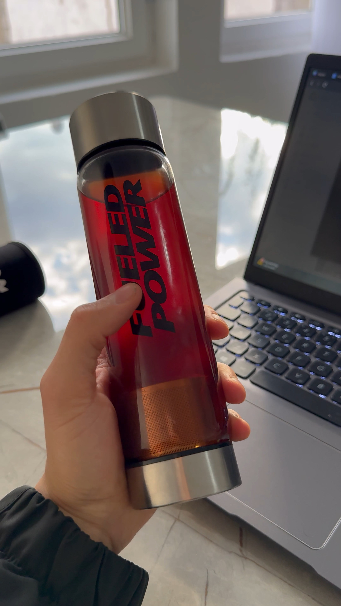A hand holds a clear bottle with red liquid and the words 'FUELED POWER' in front of a laptop.